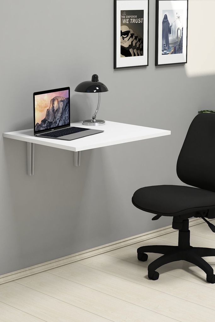 A white folding desk mounted on a gray wall, with a laptop and a lamp on it. A black office chair is in the foreground.