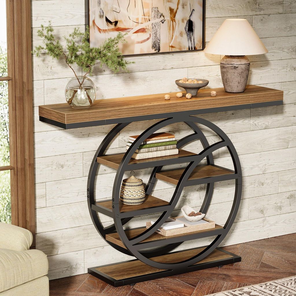 A console table with a geometric metal frame and wooden shelves is placed against a wood-paneled wall, next to a window and a sofa. Decorative items like books, a vase with greenery, a lamp, and bowls are arranged on the table and shelves.