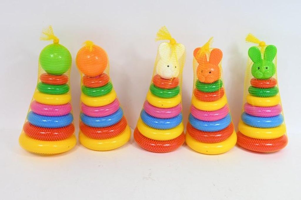 Five colorful stacking ring toys are arranged in a line against a white background. Each toy is conical in shape and made of plastic rings stacked on a central pole. The toys are individually wrapped in yellow mesh bags, tied at the top with yellow ribbon.