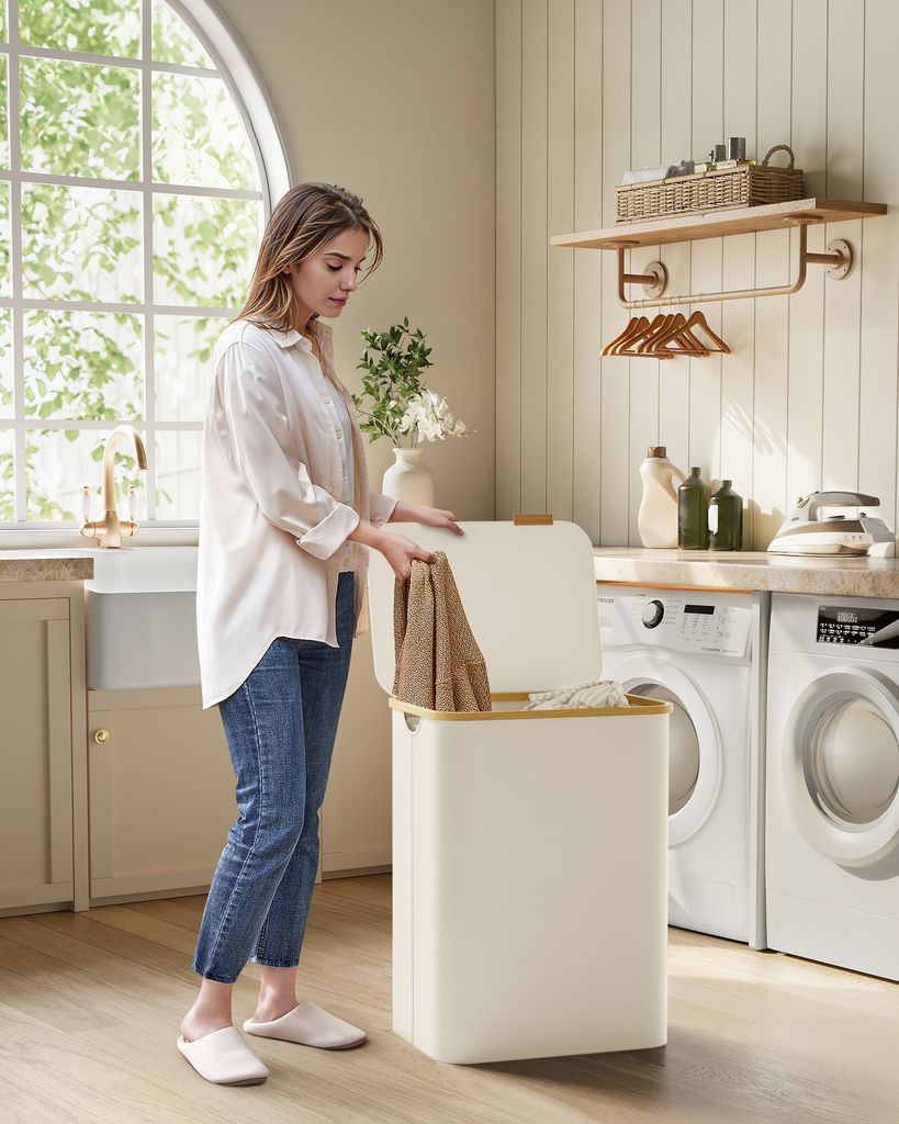 A woman is putting laundry into a large, cream-colored hamper in a laundry room.