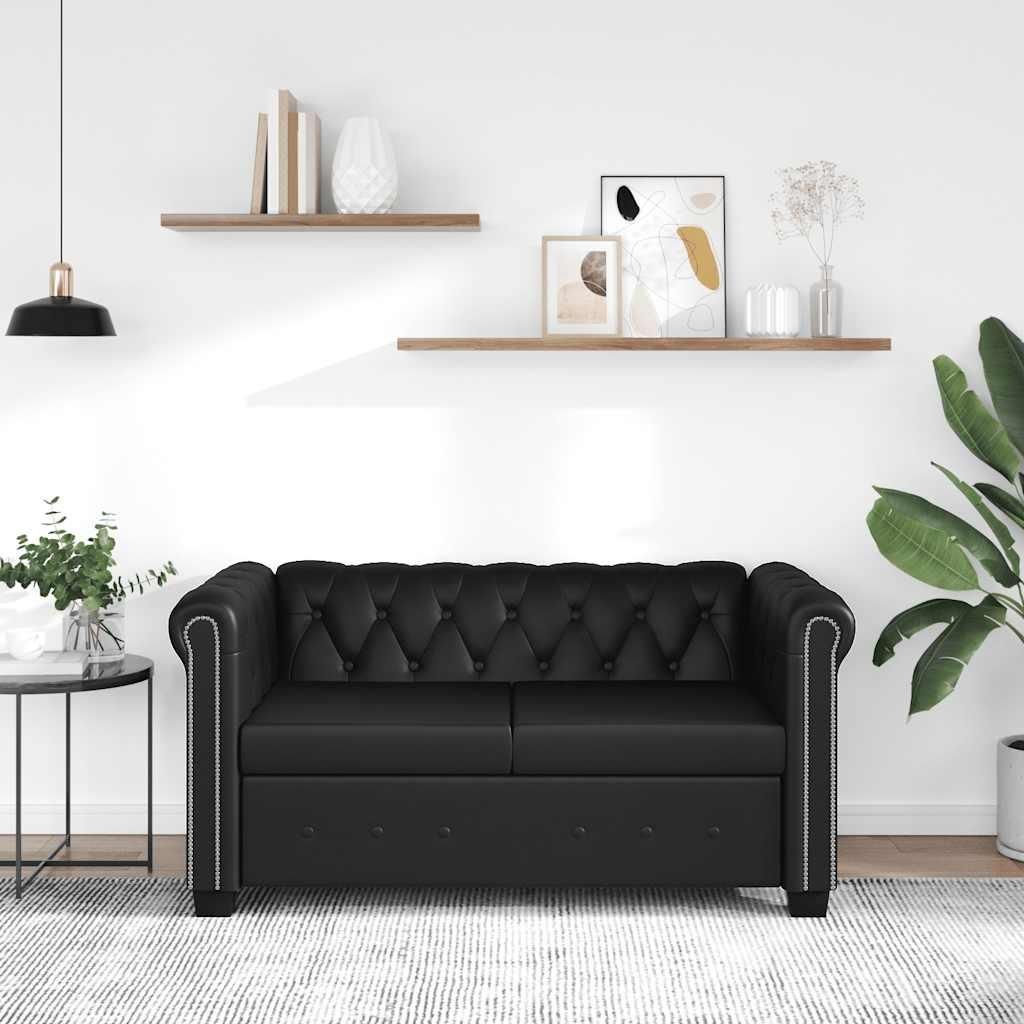 A black Chesterfield sofa is shown in a well-lit room with shelves on the wall.