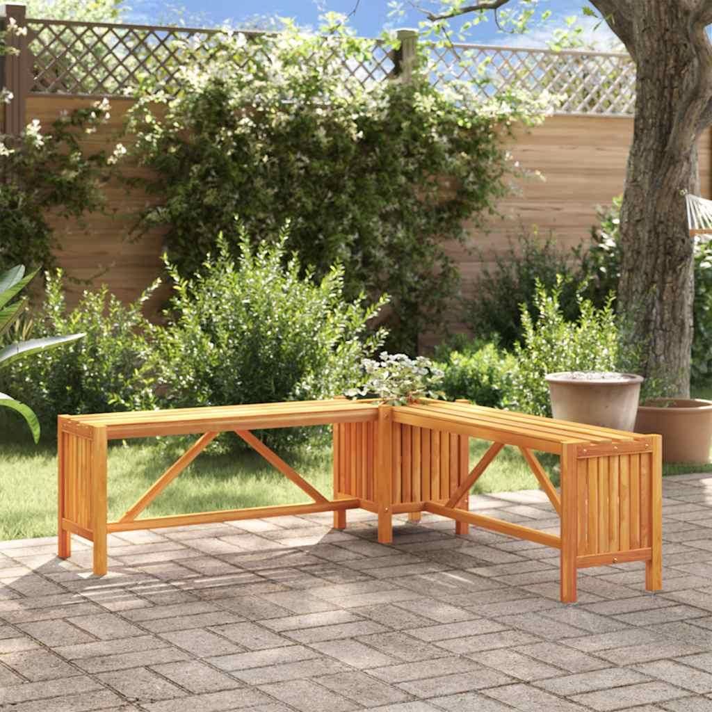 A wooden corner bench with a planter is situated on a paved patio in a garden setting, surrounded by greenery and a tree.