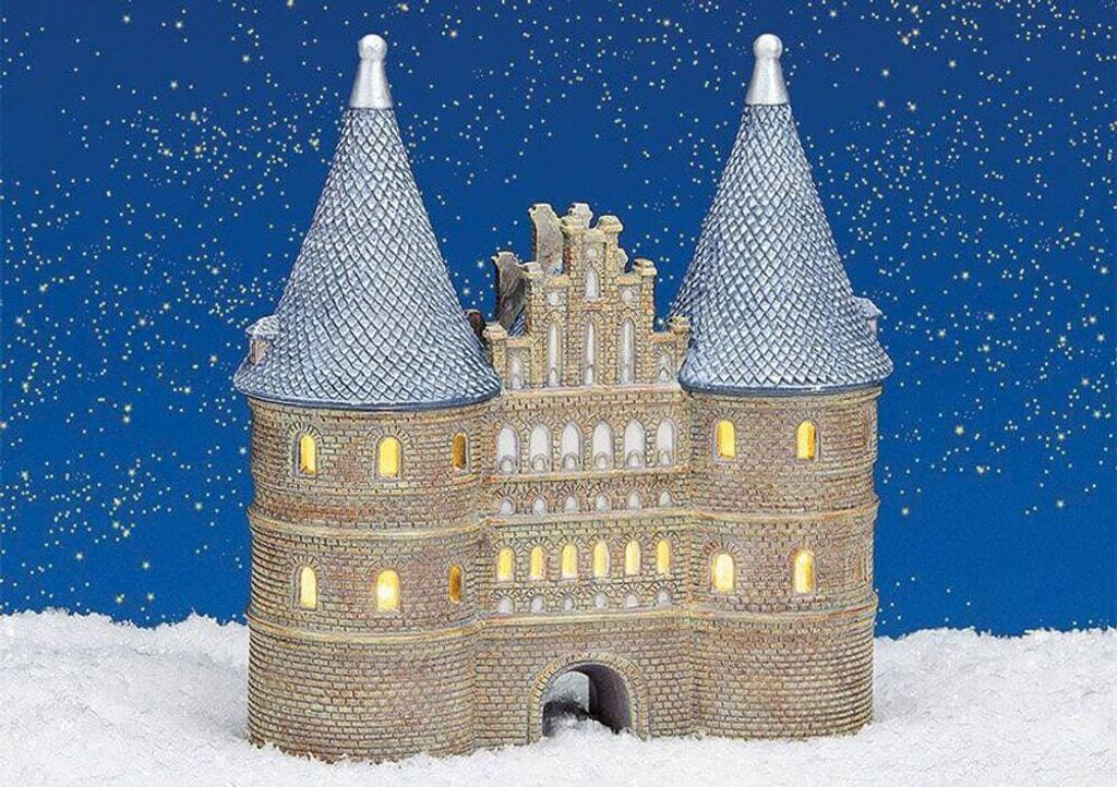 A miniature, illuminated castle-like building, likely a Christmas village decoration, sits in the snow under a starry night sky.
