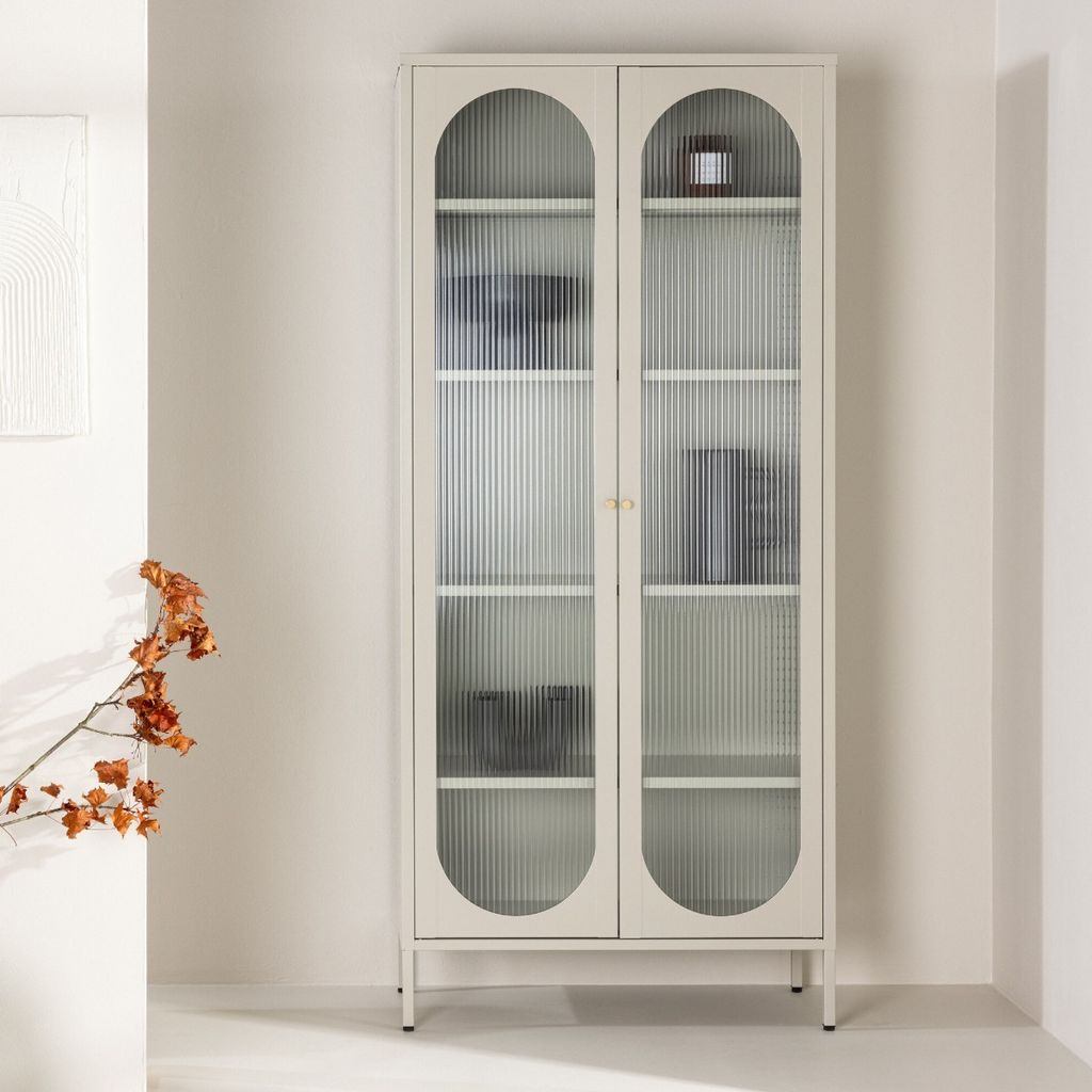 A beige display cabinet with fluted glass doors and arched tops, standing in a minimalist room with a branch of dried flowers to the left.