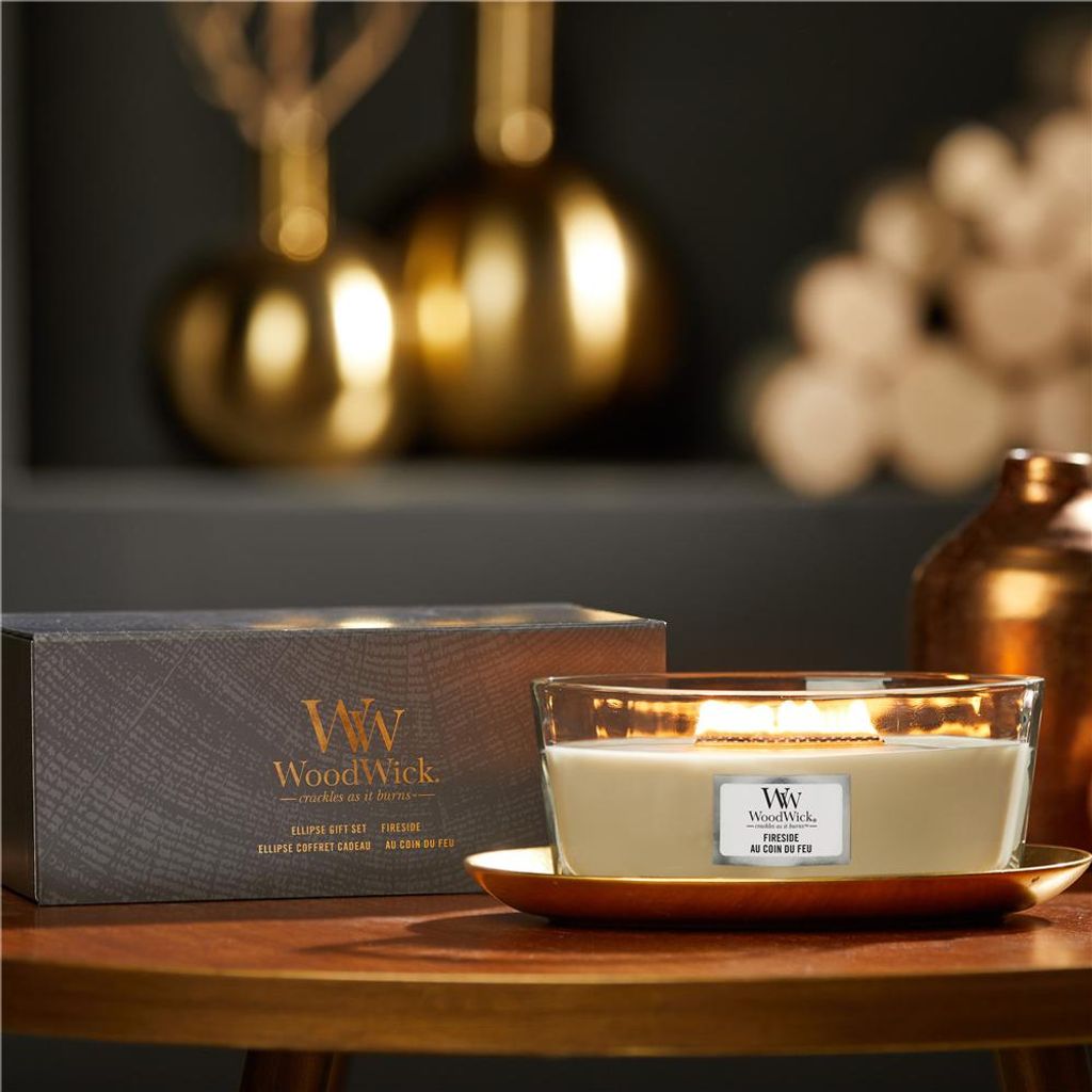 A WoodWick candle and its packaging are displayed on a wooden table with decorative golden objects in the background.