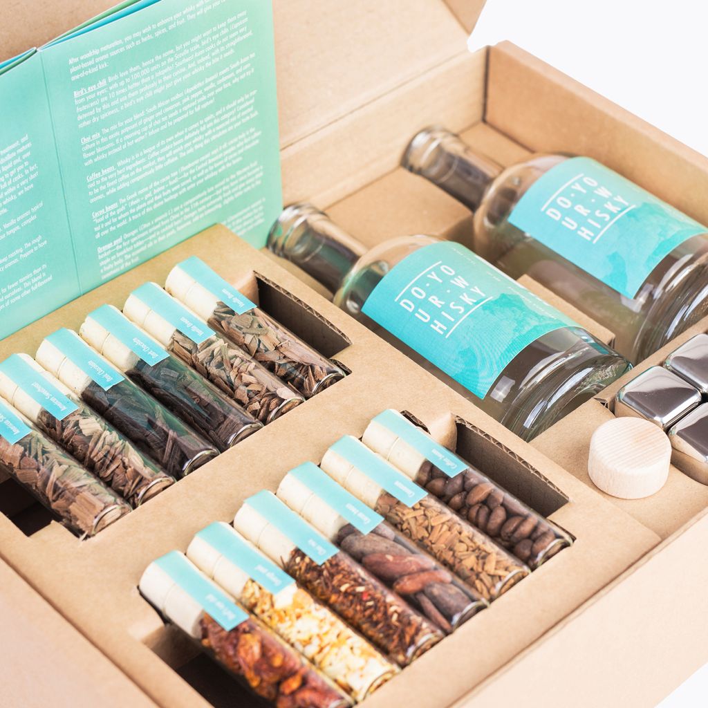 A whiskey making kit laid out in a cardboard box, featuring small bottles of ingredients, two empty glass bottles, and ice cubes.