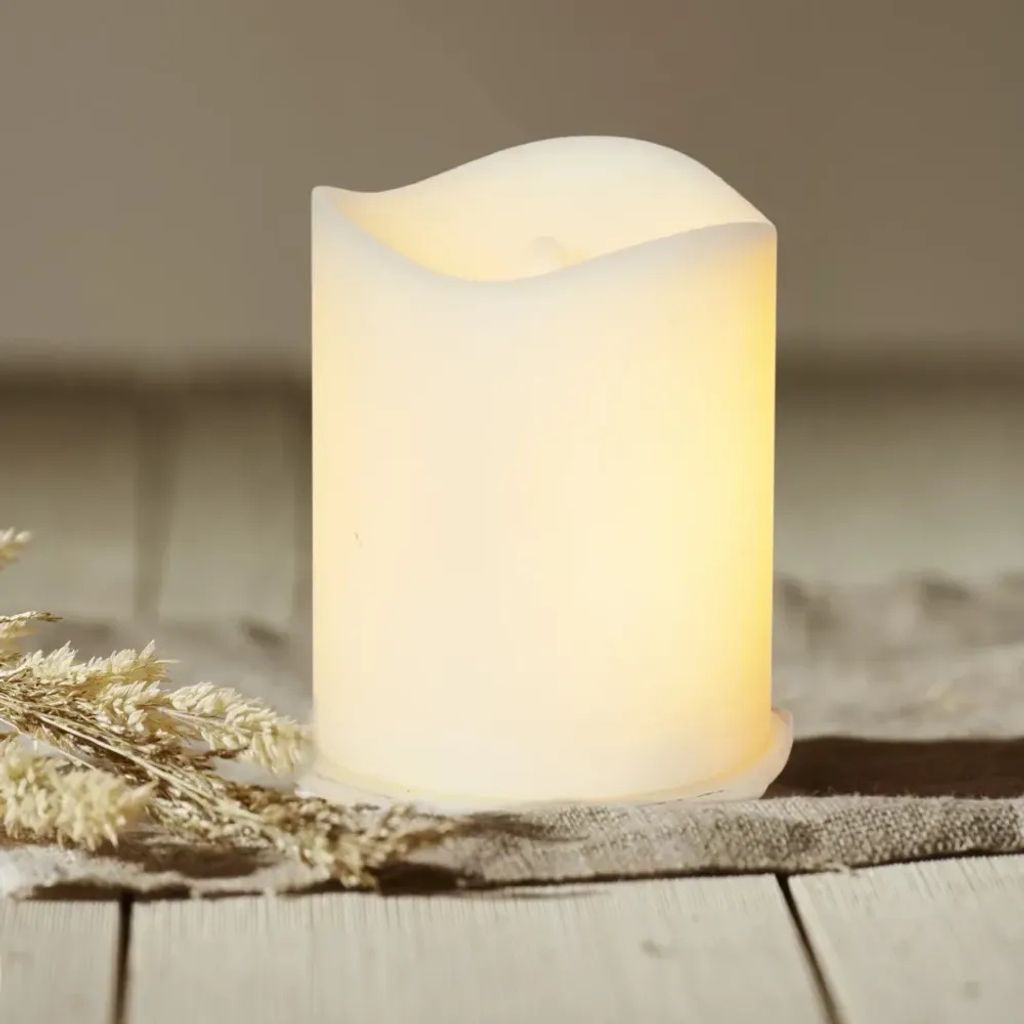 A white LED candle with a flame-like top sits on a textured fabric runner, next to some dried wheat stalks, on a wooden surface.