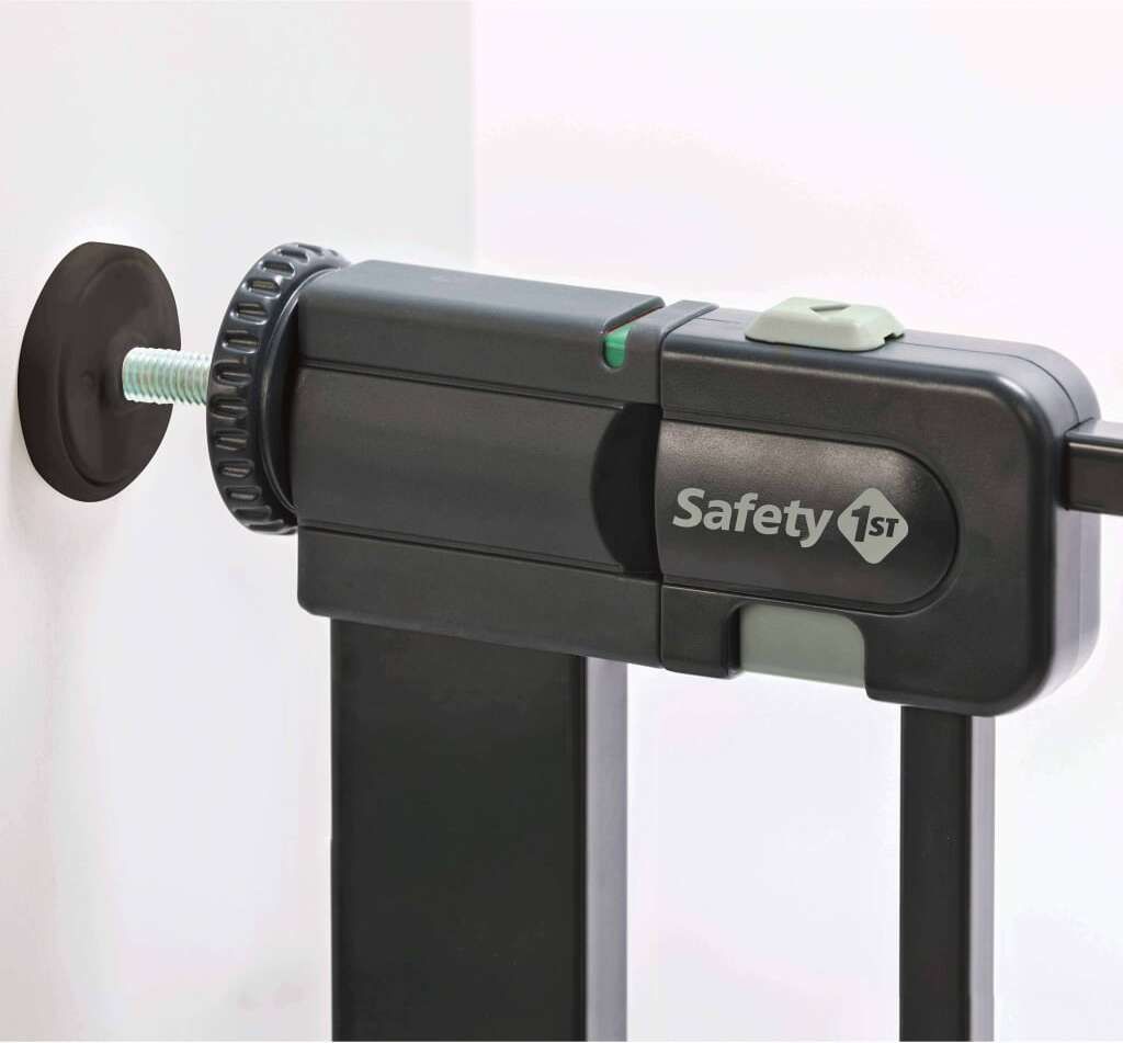 A close-up view of a black metal safety gate mechanism, showing the mounting hardware and the 'Safety 1st' logo.