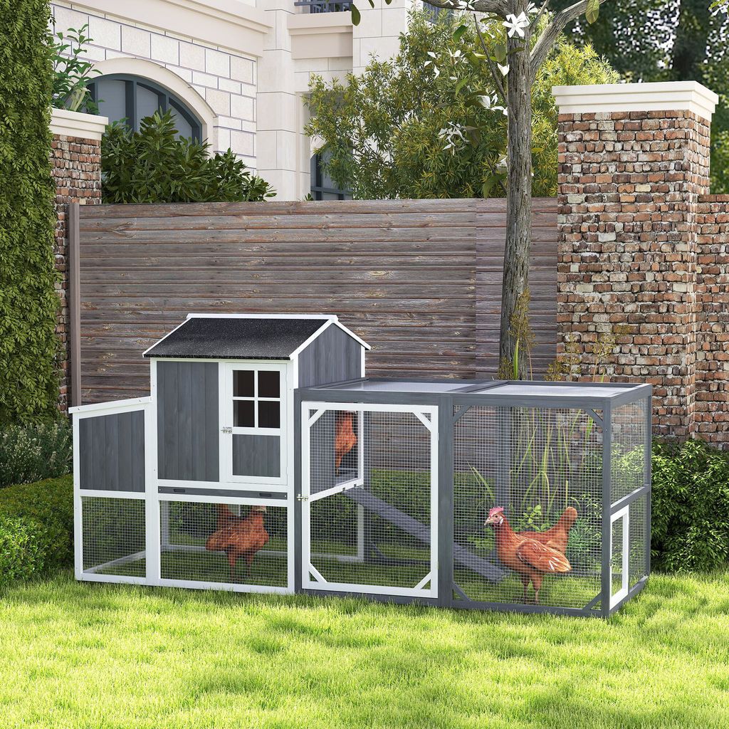 A wooden chicken coop with an attached run is set up on a grassy lawn in front of a wooden fence and a brick wall. Two chickens are visible inside the coop and run.