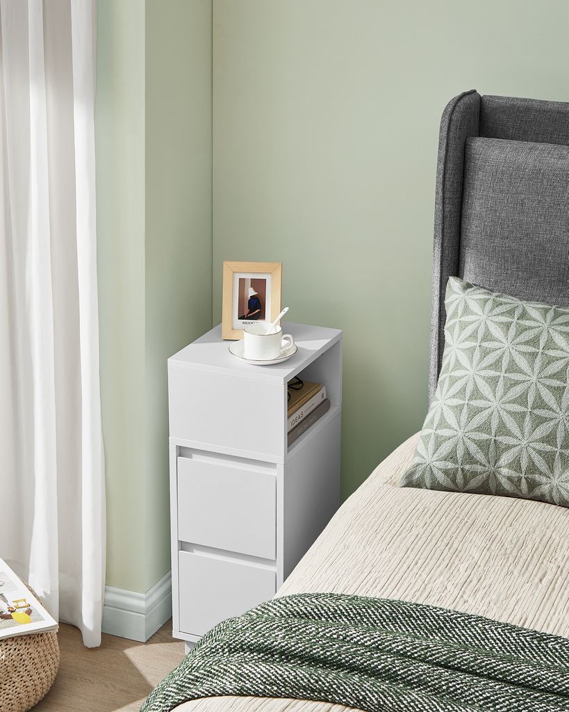 A white bedside table with drawers next to a bed with a grey headboard and a patterned pillow.