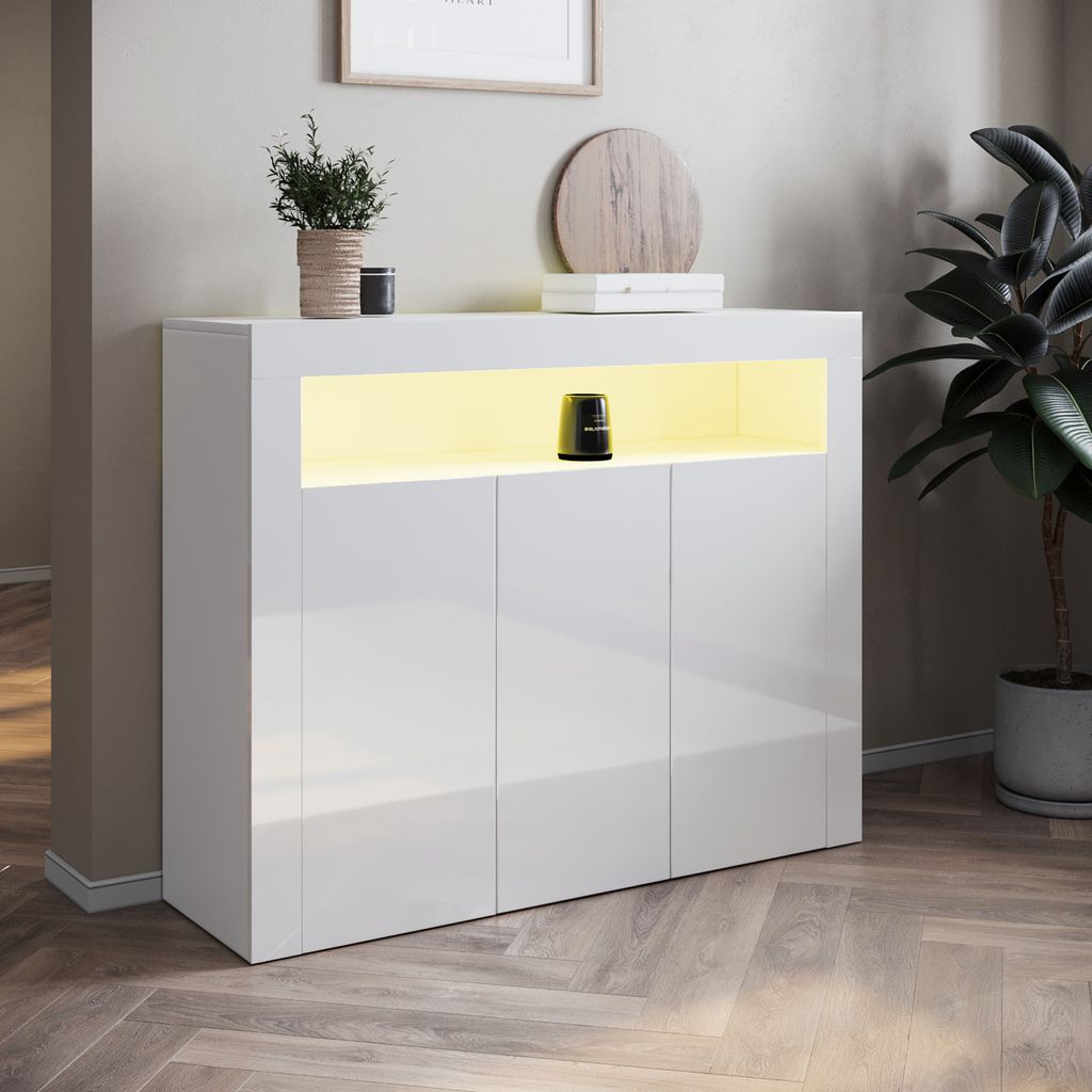 A white high-gloss sideboard with LED lighting is displayed in a room with a plant and artwork.