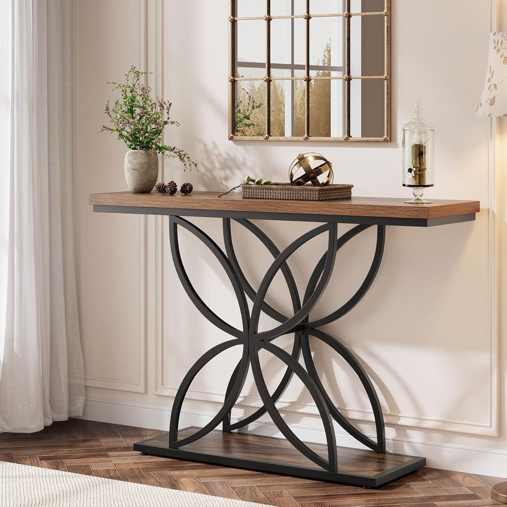 A console table with decorative items is placed against a wall with molding and a mirror above it. A window with sheer curtains is to the left.