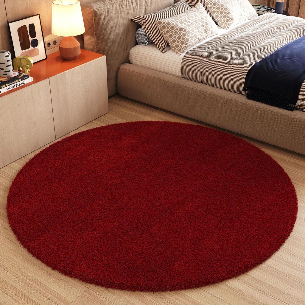 A dark red round rug is placed on a light wood floor in front of a bed and nightstand.