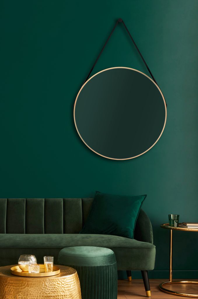 A round mirror with a gold frame hangs on a dark green wall above a green velvet sofa. A green ottoman and a gold coffee table with drinks are in the foreground.