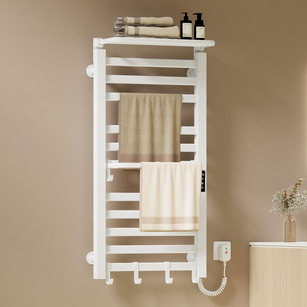 A white electric towel warmer mounted on a light brown wall, with two towels draped over it. There are also some bottles and rolled towels on a shelf above the warmer, and a small vase with dried flowers on a cabinet to the right.