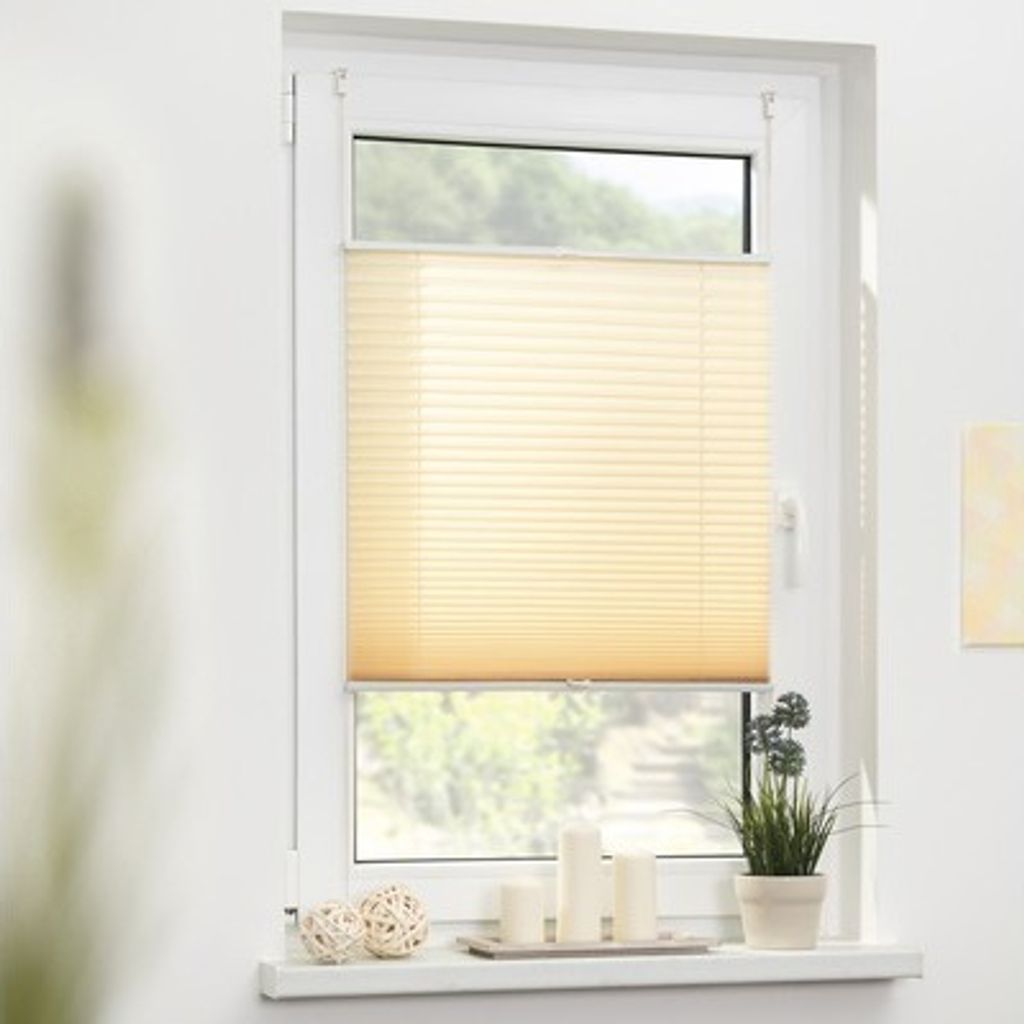 A cream-colored pleated blind is installed on a white window frame, with a view of greenery outside. Decorative items like balls and candles are on the windowsill.