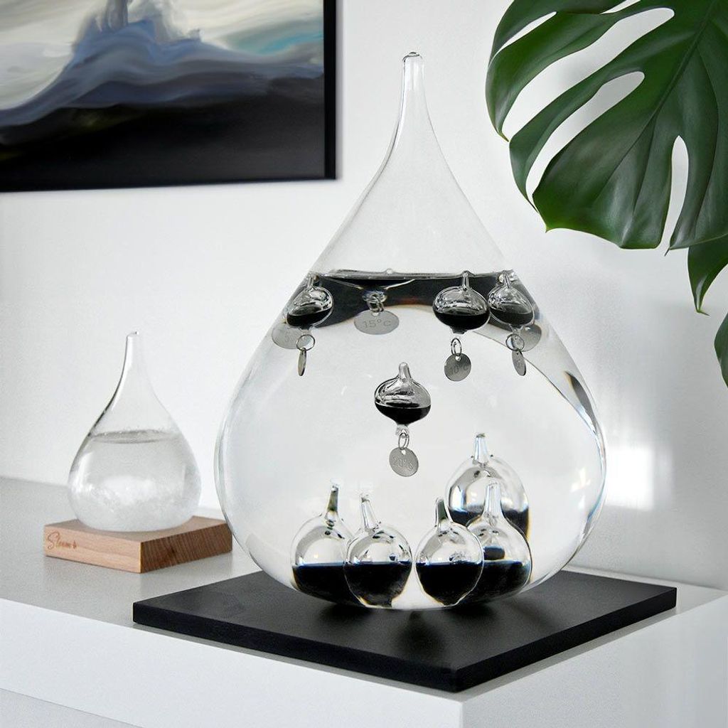 A Galileo thermometer is displayed on a shelf next to a smaller glass container, with a plant and artwork in the background.