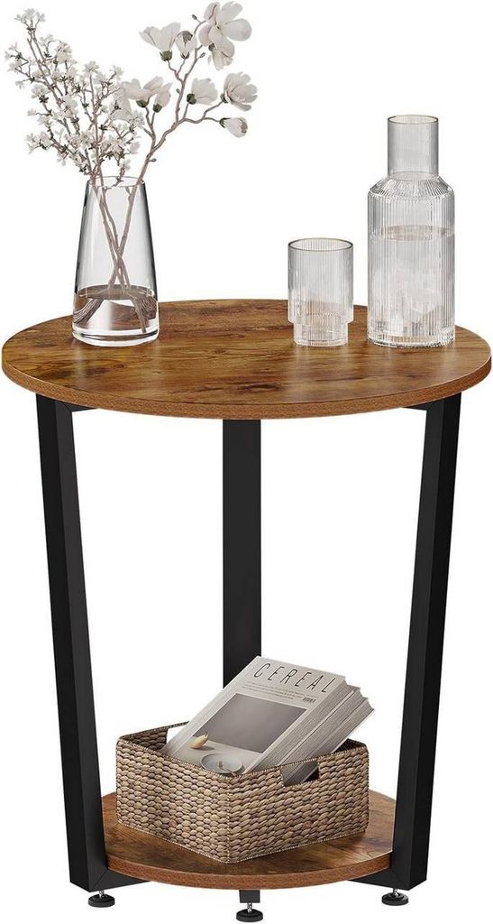 A round side table with a wood-look top and black metal legs, featuring a vase with flowers, a glass, and a bottle on top, and a basket with magazines on the lower shelf.