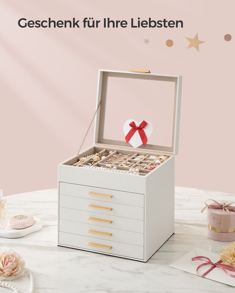 A white jewelry box with multiple drawers and a glass lid is open, displaying various jewelry items. A small heart-shaped decoration with a red ribbon is attached to the inside of the lid. The box sits on a marble-like surface, with decorative elements lik