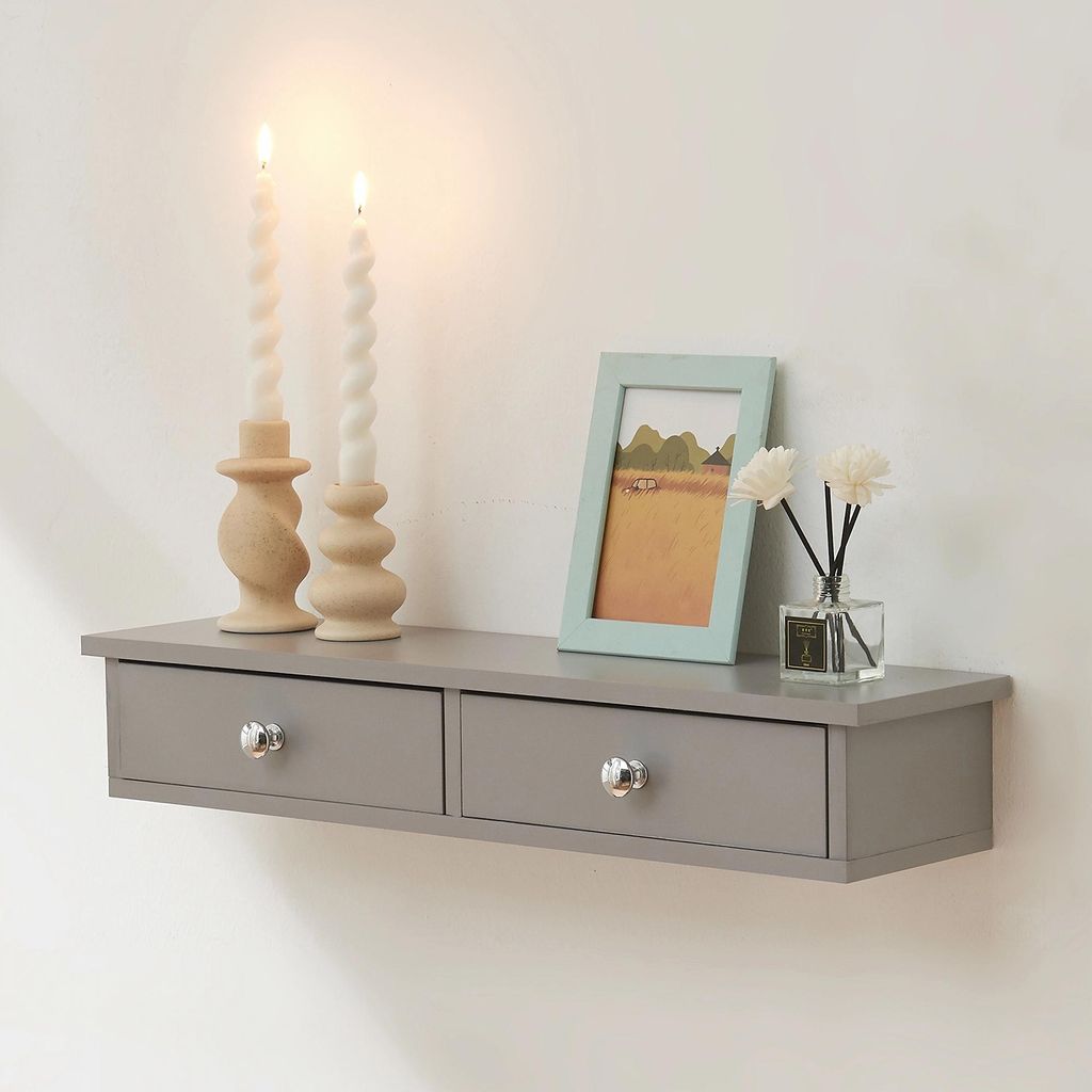A gray floating shelf with two drawers is mounted on a white wall. It is decorated with two white twisted candles on wooden candle holders, a framed picture of a landscape, and a small glass bottle with reeds.