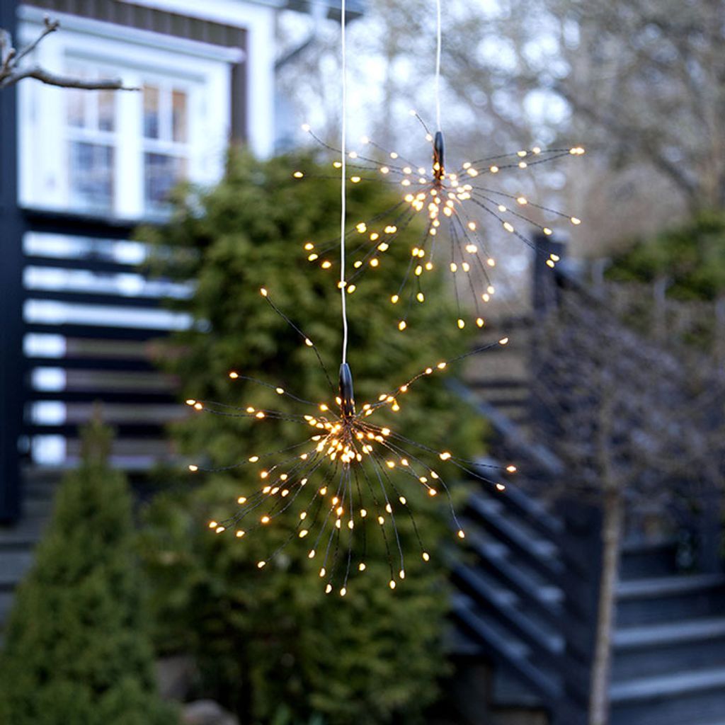 Two firework-shaped solar lights hanging outdoors in front of a house and trees.