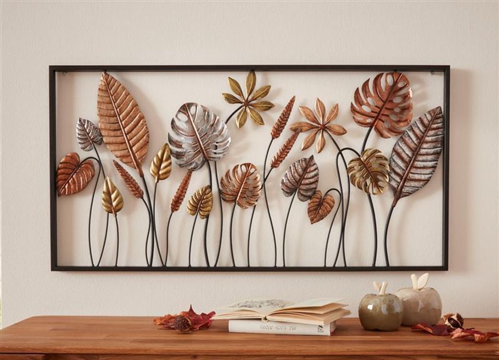 A metal wall art piece featuring various leaf designs is displayed on a wall above a wooden table with books and decorative apples.