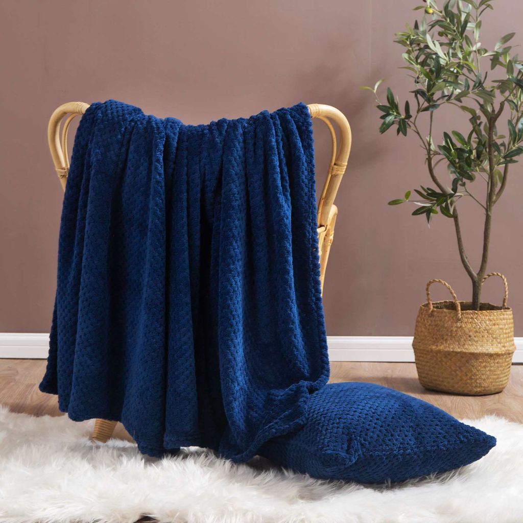 A navy blue blanket draped over a wicker chair, with a matching pillow on a white rug and a potted plant in the background.