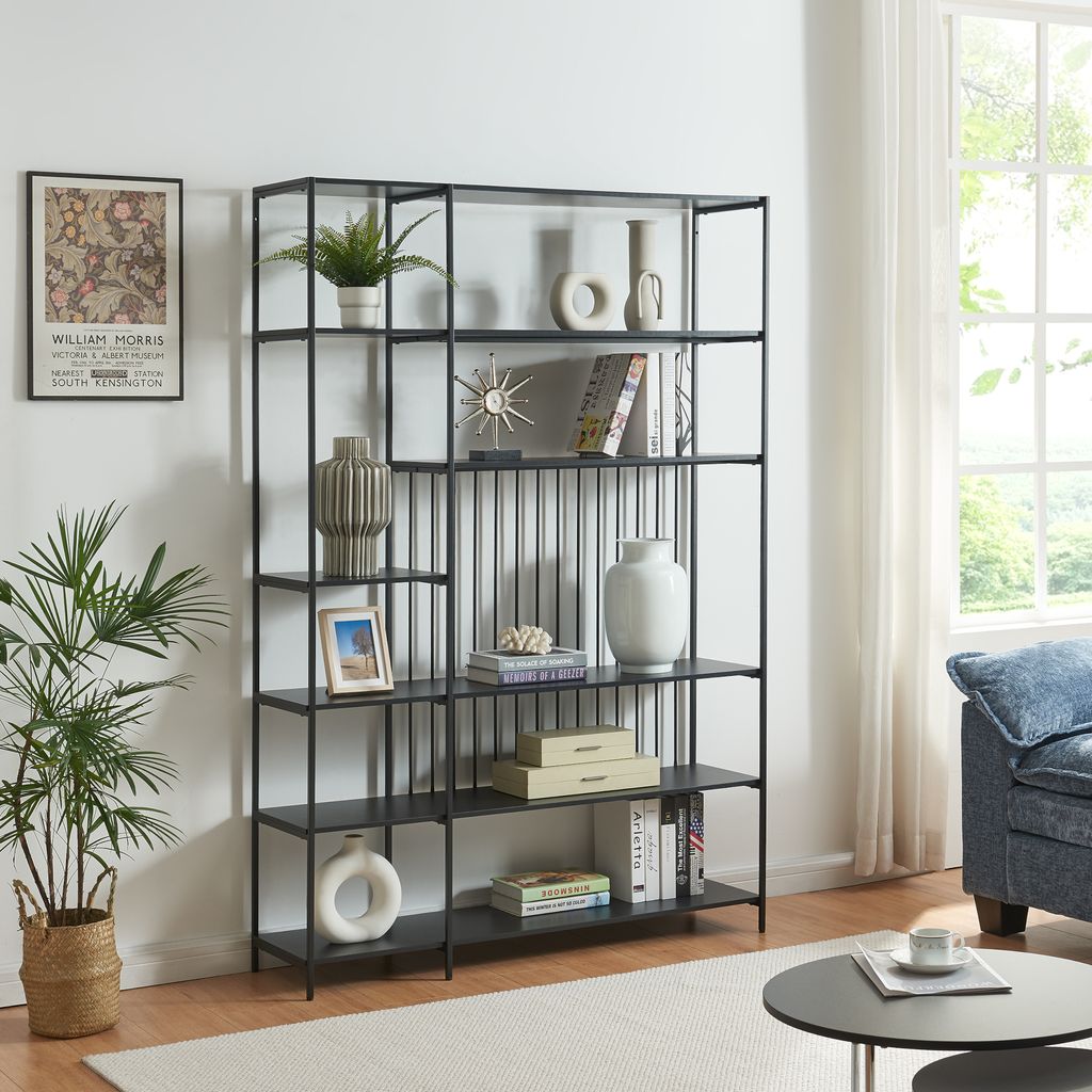 A black metal bookshelf filled with decorative items and books, placed in a living room setting next to a window and a blue sofa.