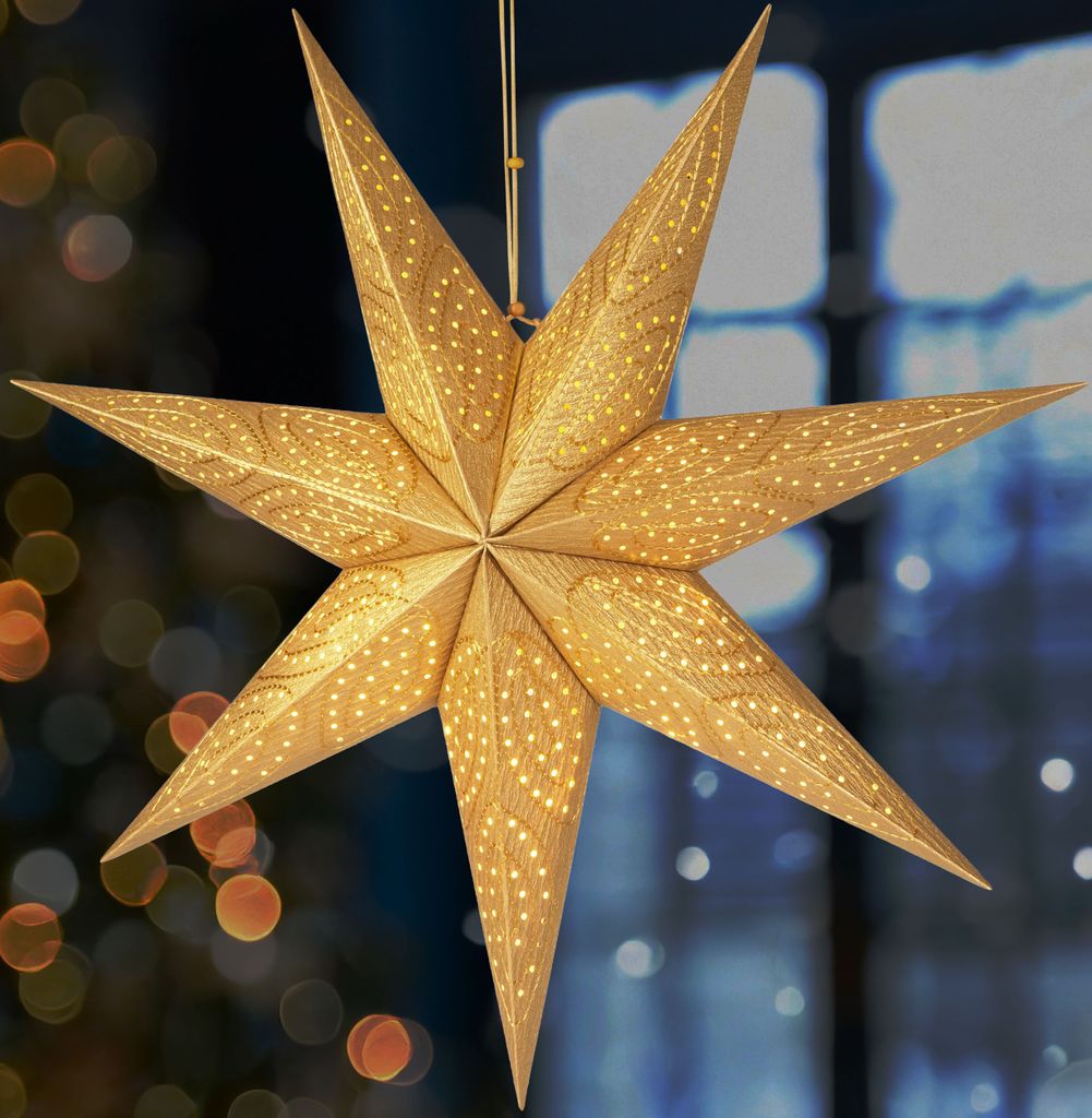A golden, 3D paper star decoration hangs in front of a blurred background of lights and a window.