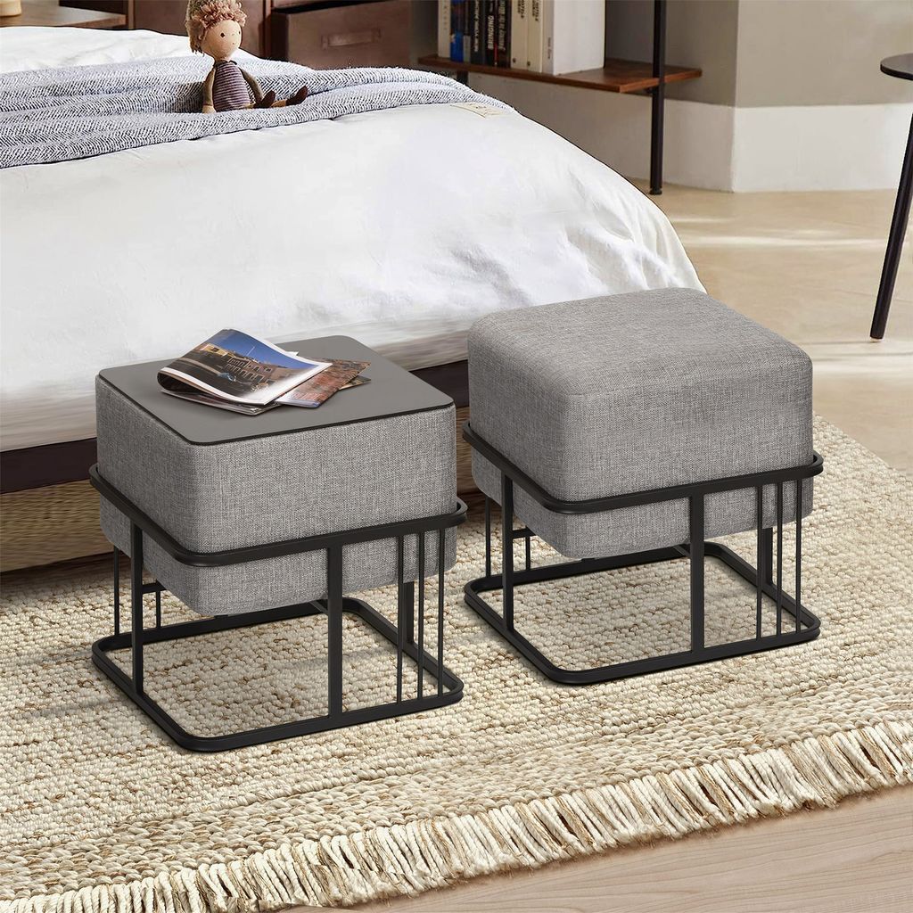 Two gray upholstered ottomans with black metal frames are placed on a textured rug next to a bed.