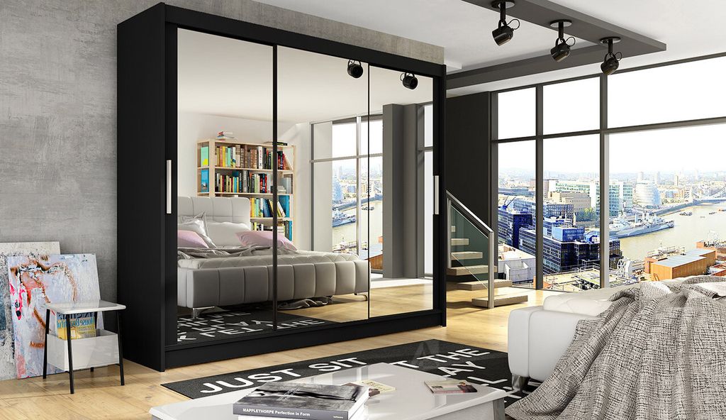 A bedroom with a large black wardrobe with mirrored doors, a bed, and a view of a city from a large window.