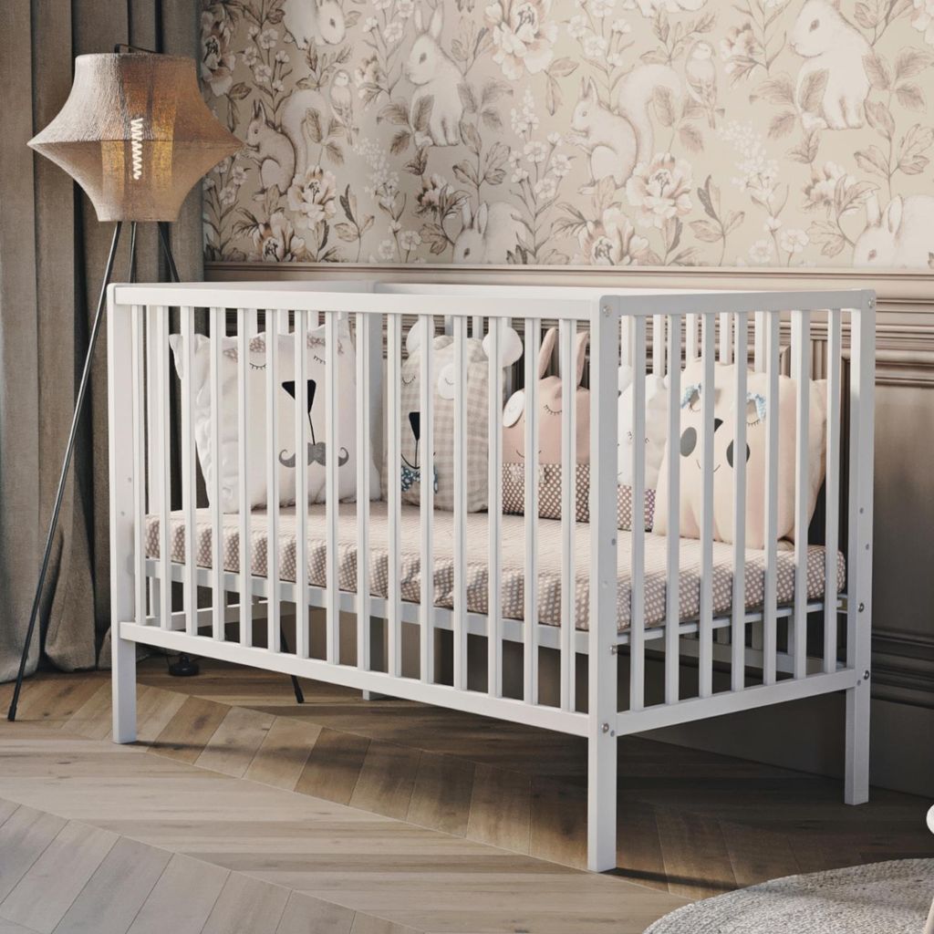 A white baby crib with decorative pillows inside, set in a nursery with patterned wallpaper and a floor lamp.