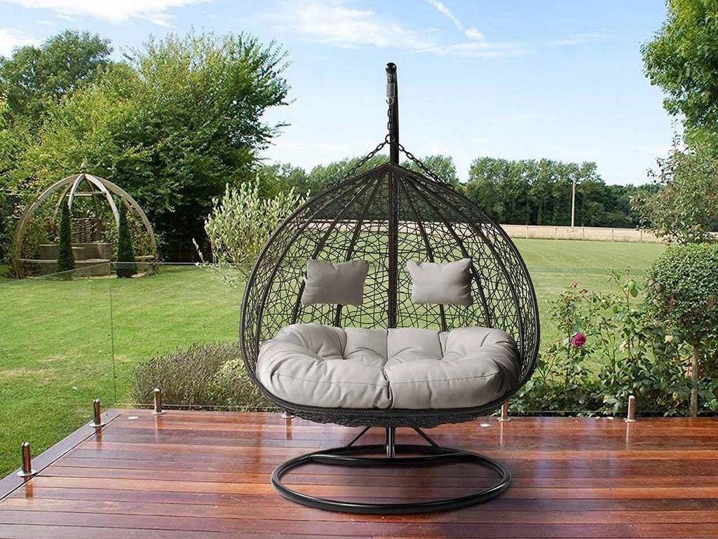 A hanging egg chair for two people is placed on a wooden deck in a garden setting.
