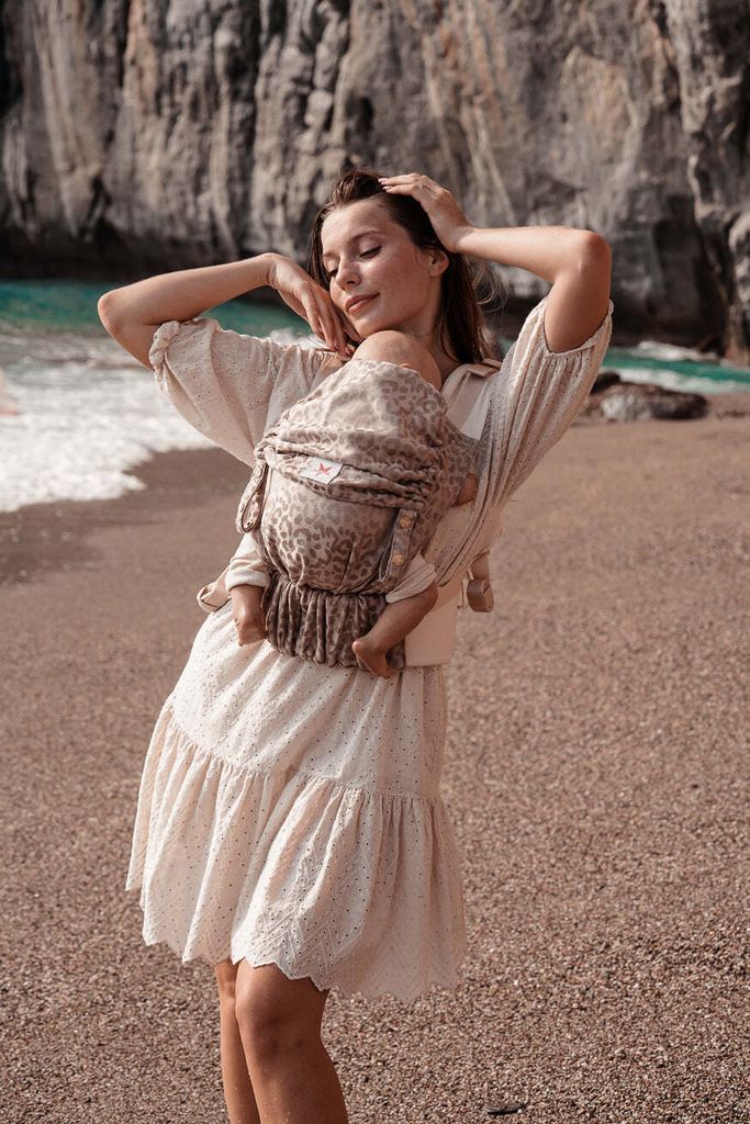 A woman is carrying a baby in a sling on a beach with cliffs and the ocean in the background.