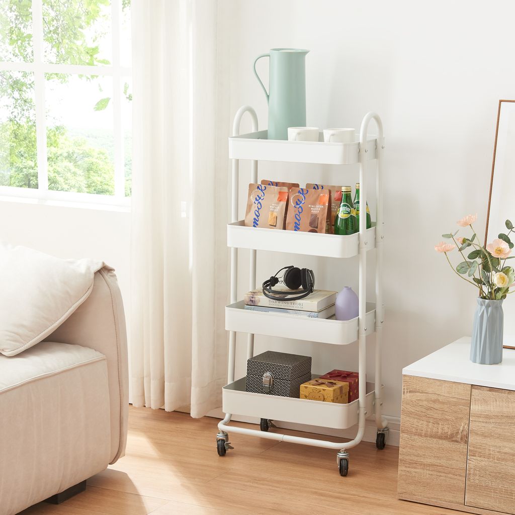 A white rolling cart with four shelves filled with various items, placed next to a sofa and a cabinet with flowers.