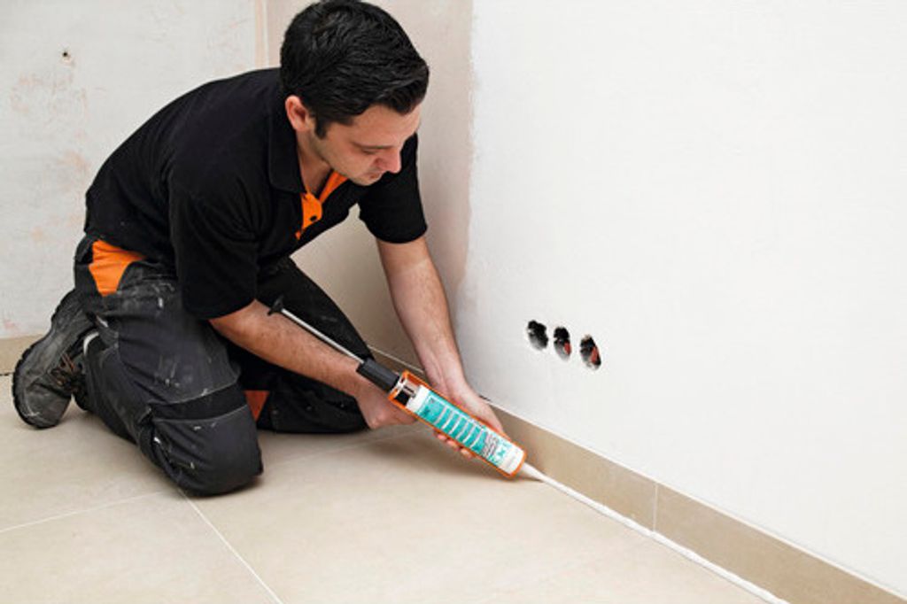 A man is kneeling and applying sealant to the base of a wall.