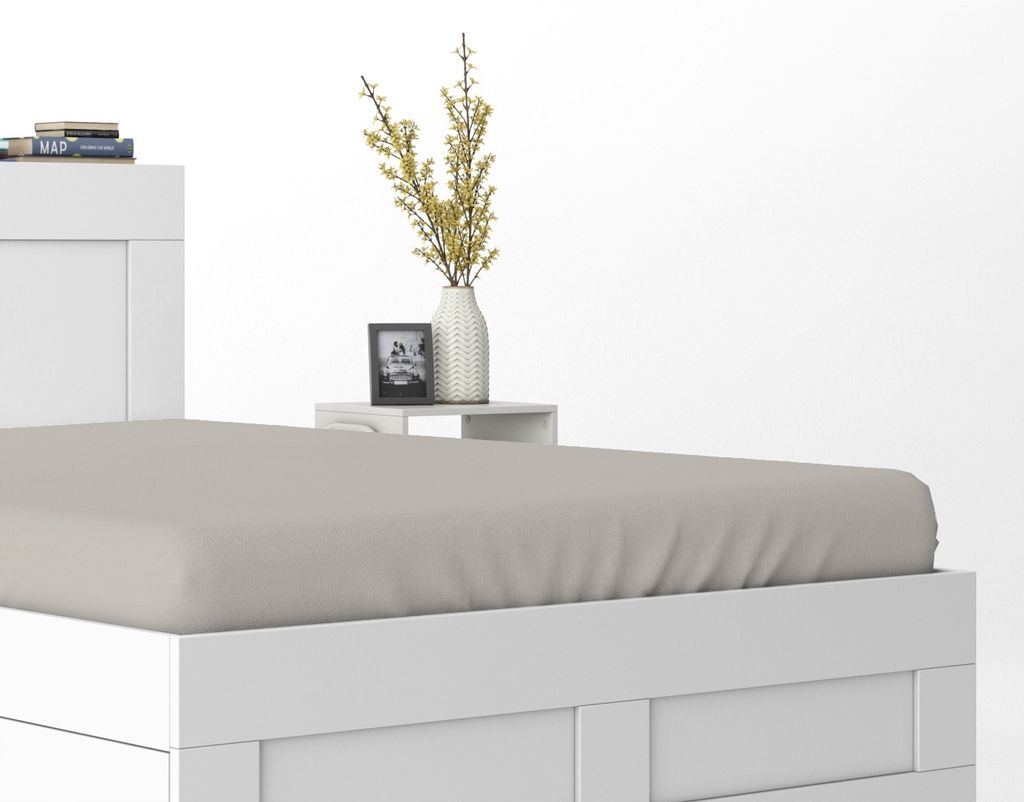 A close-up view of a white bed frame with a beige fitted sheet, next to a small white nightstand with a vase of yellow flowers and a framed picture.