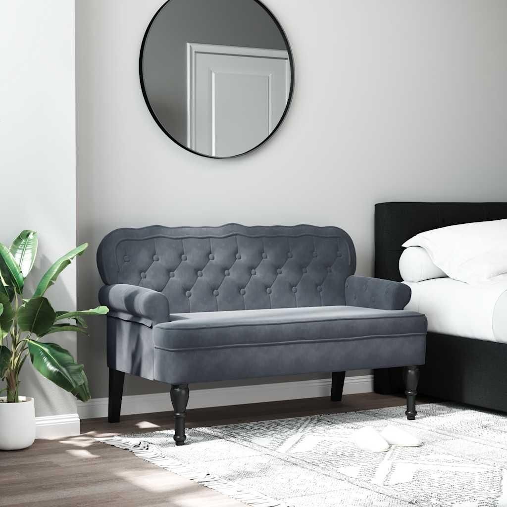 A dark gray velvet bench with a tufted backrest is placed in a bedroom setting next to a bed. A large round mirror hangs on the wall behind the bench, and a potted plant is visible to the left.