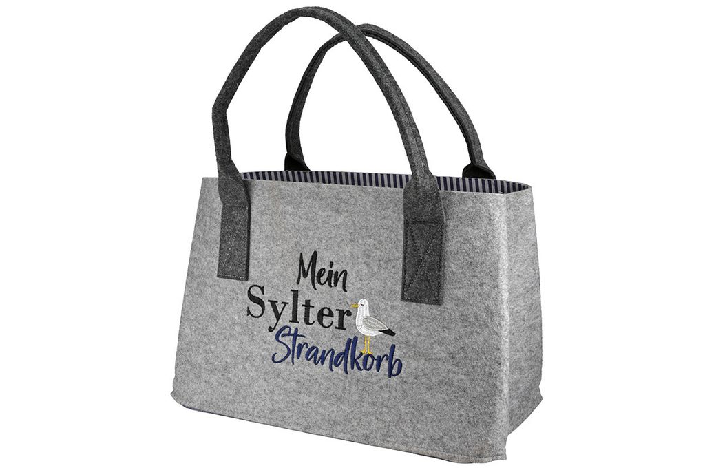 A light gray felt tote bag with dark gray handles and a navy and white striped lining. The bag has an embroidered seagull and text that reads "Mein Sylter Strandkorb".