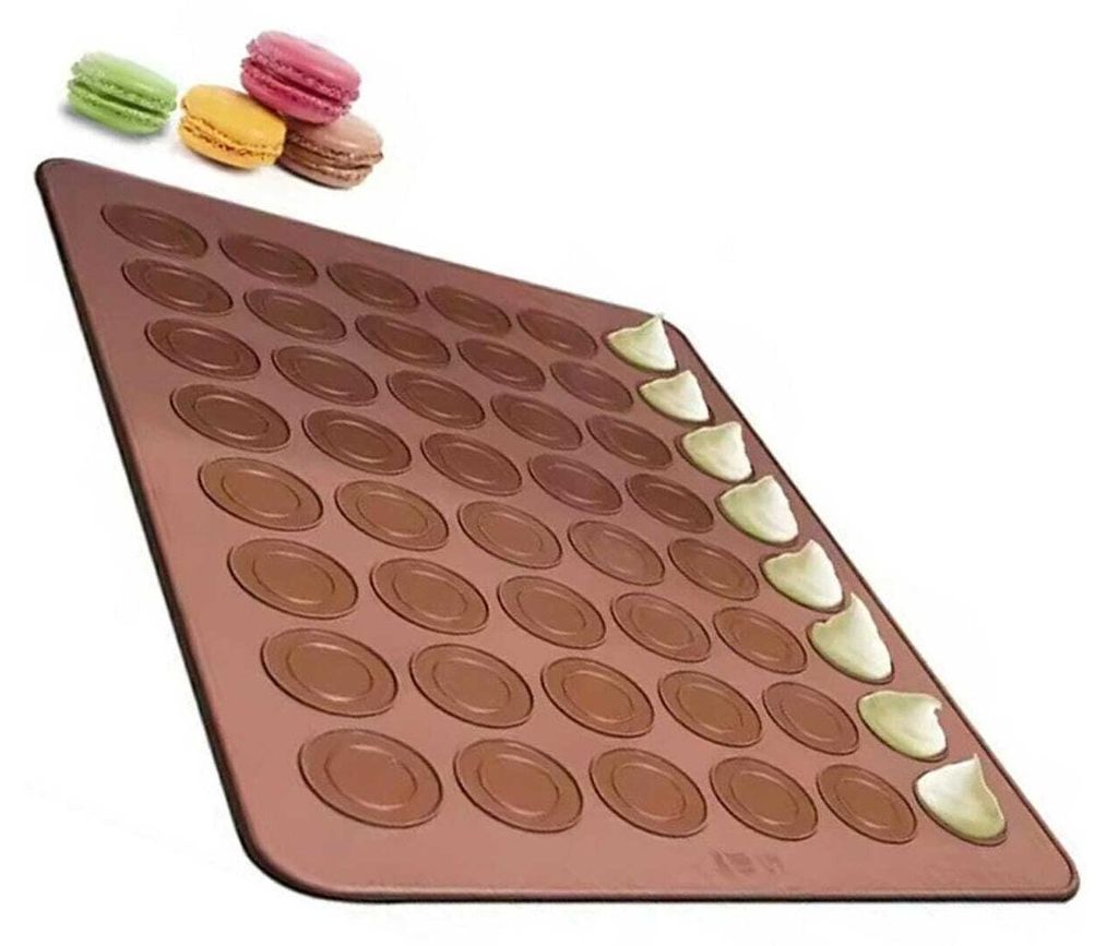 A silicone baking mat with circular indentations for making macarons, with a few macarons shown at the top left and some filling being piped onto the mat.