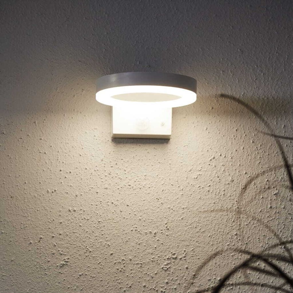 A modern, circular wall light fixture is mounted on a textured, light-colored wall. The light is illuminated, casting a soft glow and a shadow of a plant on the wall.