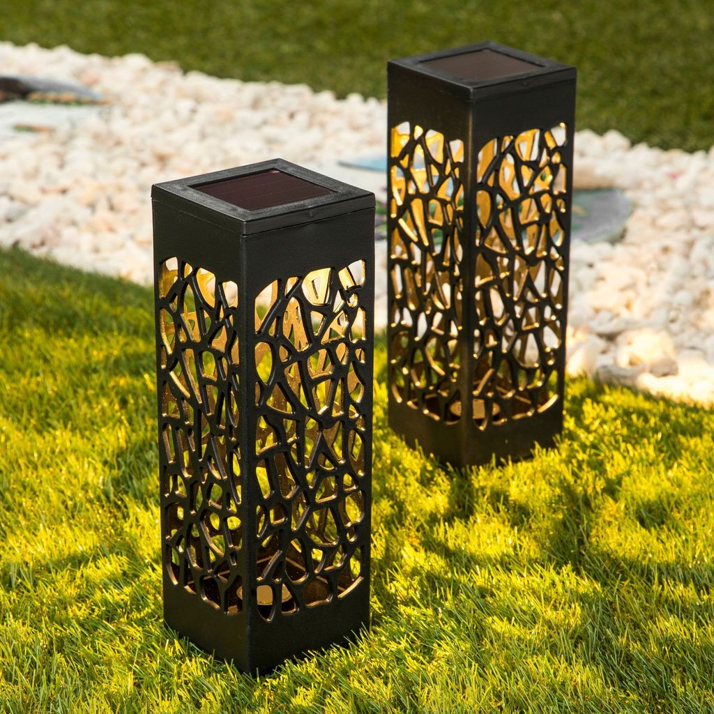 Two black solar garden lights with intricate cutouts are placed on a green lawn next to a path of white gravel.