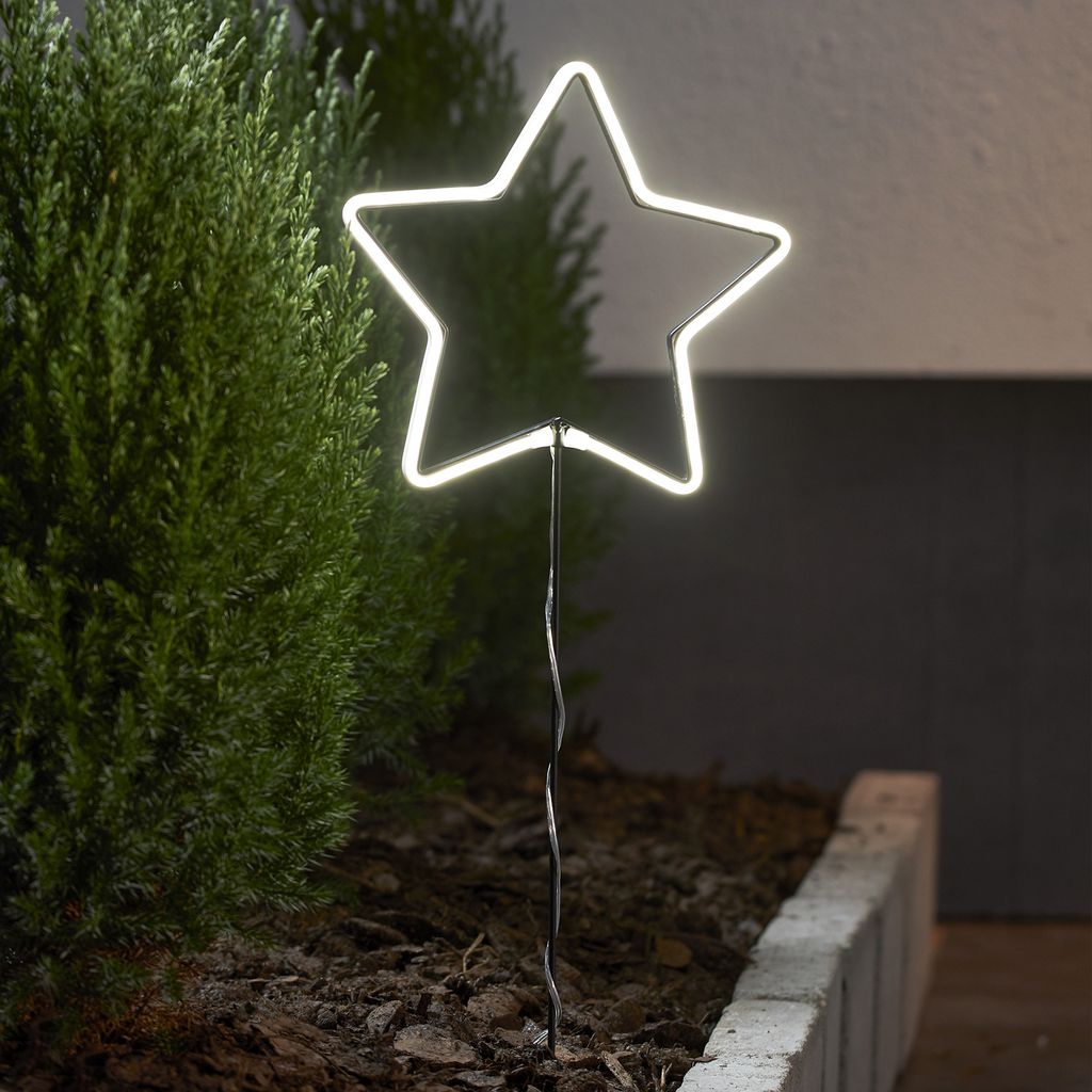A white neon star light is planted in the ground next to a green bush.