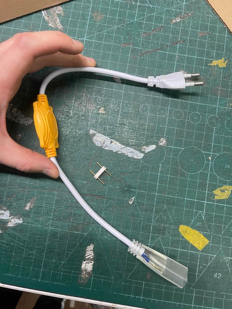 A hand holding a white power adapter with a yellow connector, resting on a green cutting mat.