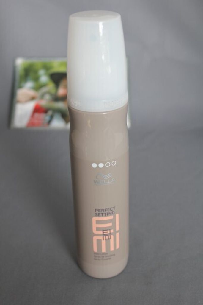 A bottle of hairspray is placed on a gray surface, with a blurred magazine in the background.