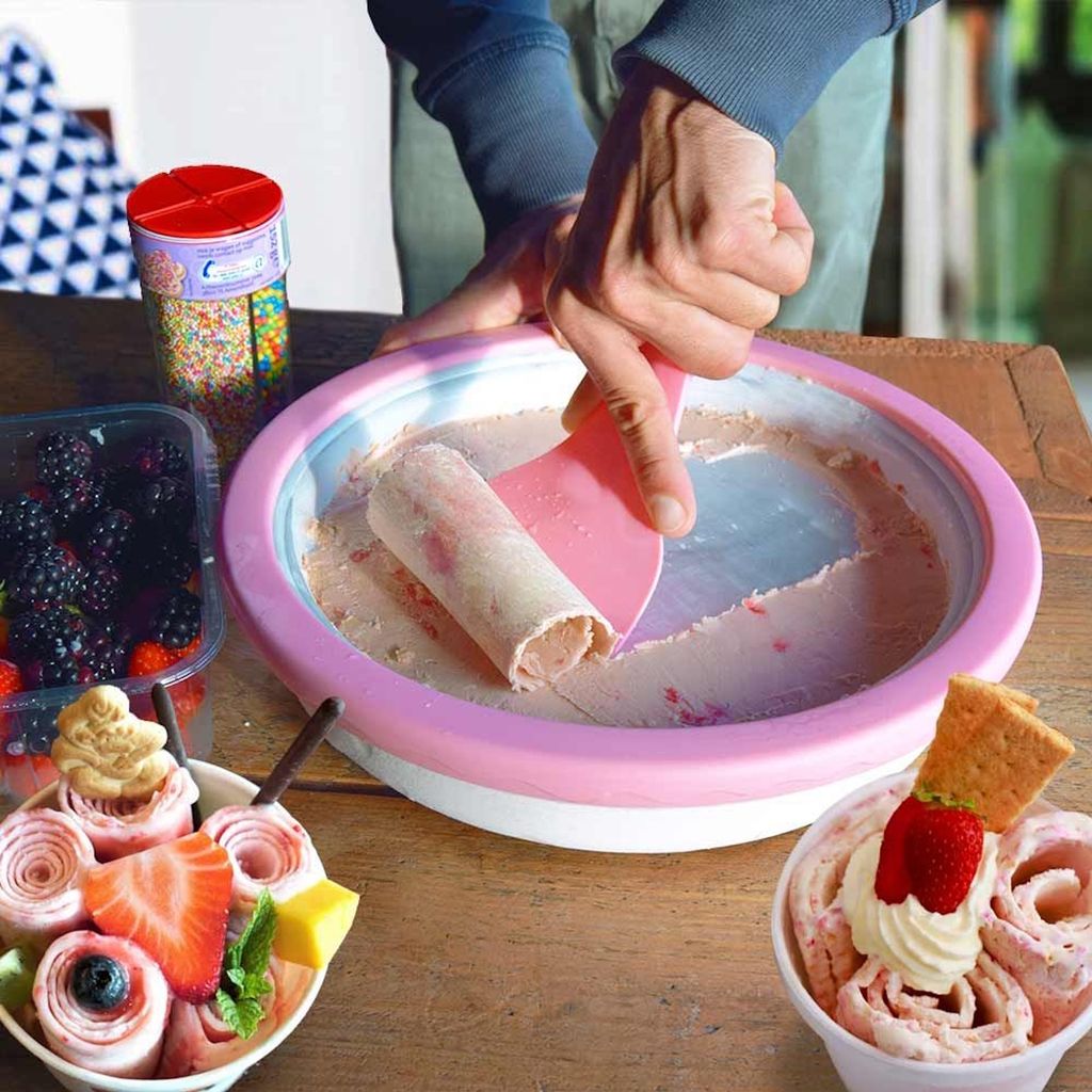 A person is making rolled ice cream on a frozen plate, with berries and finished ice cream cups nearby.