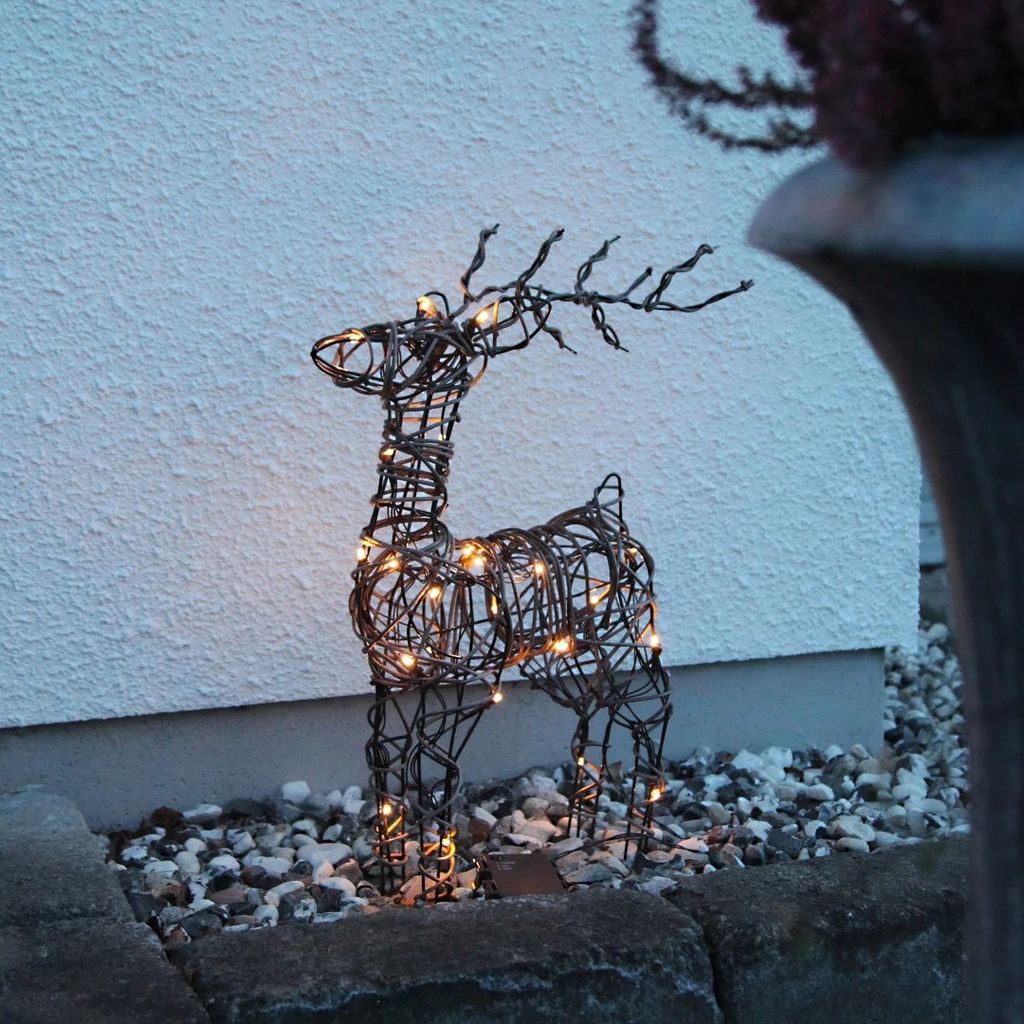 A lighted wire reindeer decoration sits on gravel in front of a white wall.