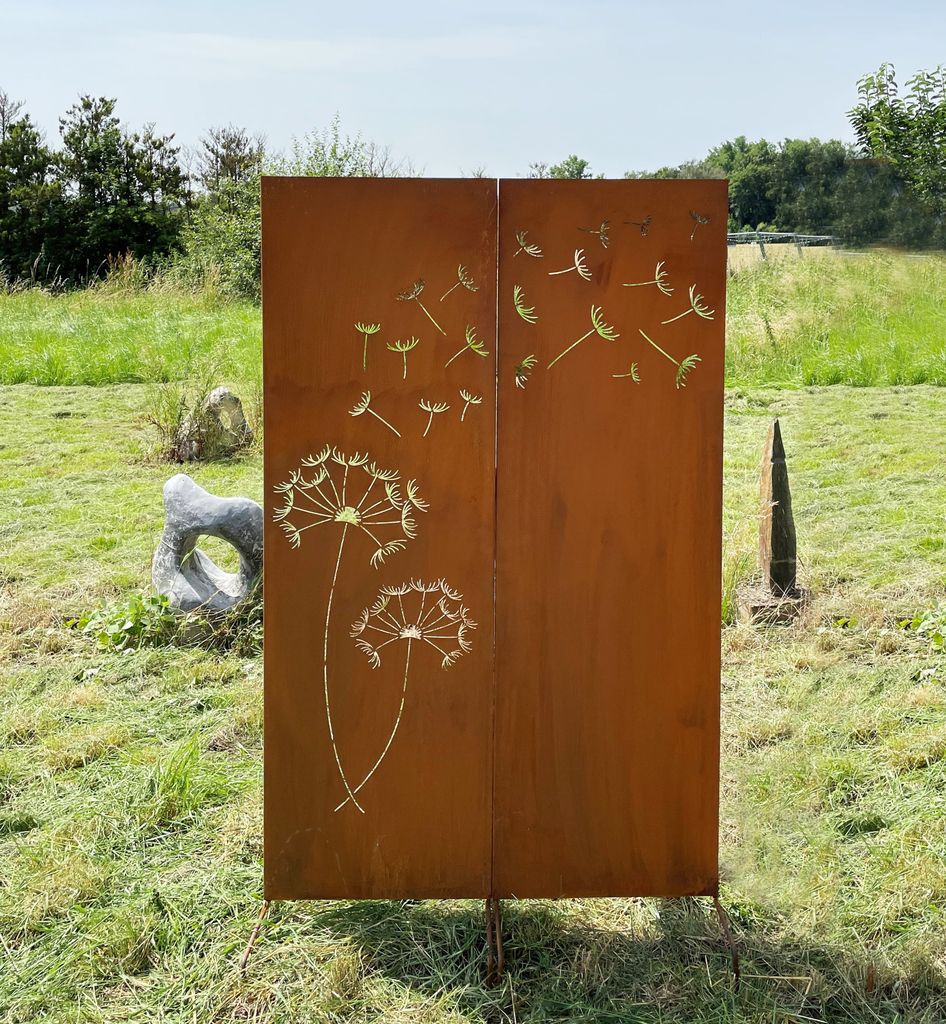 A rusty metal garden screen with a dandelion motif is placed in a grassy outdoor area with trees and sculptures in the background.