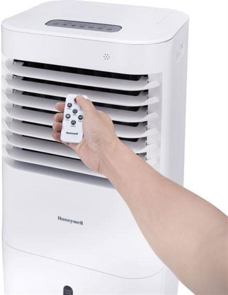 A hand holding a remote control in front of a Honeywell air cooler.