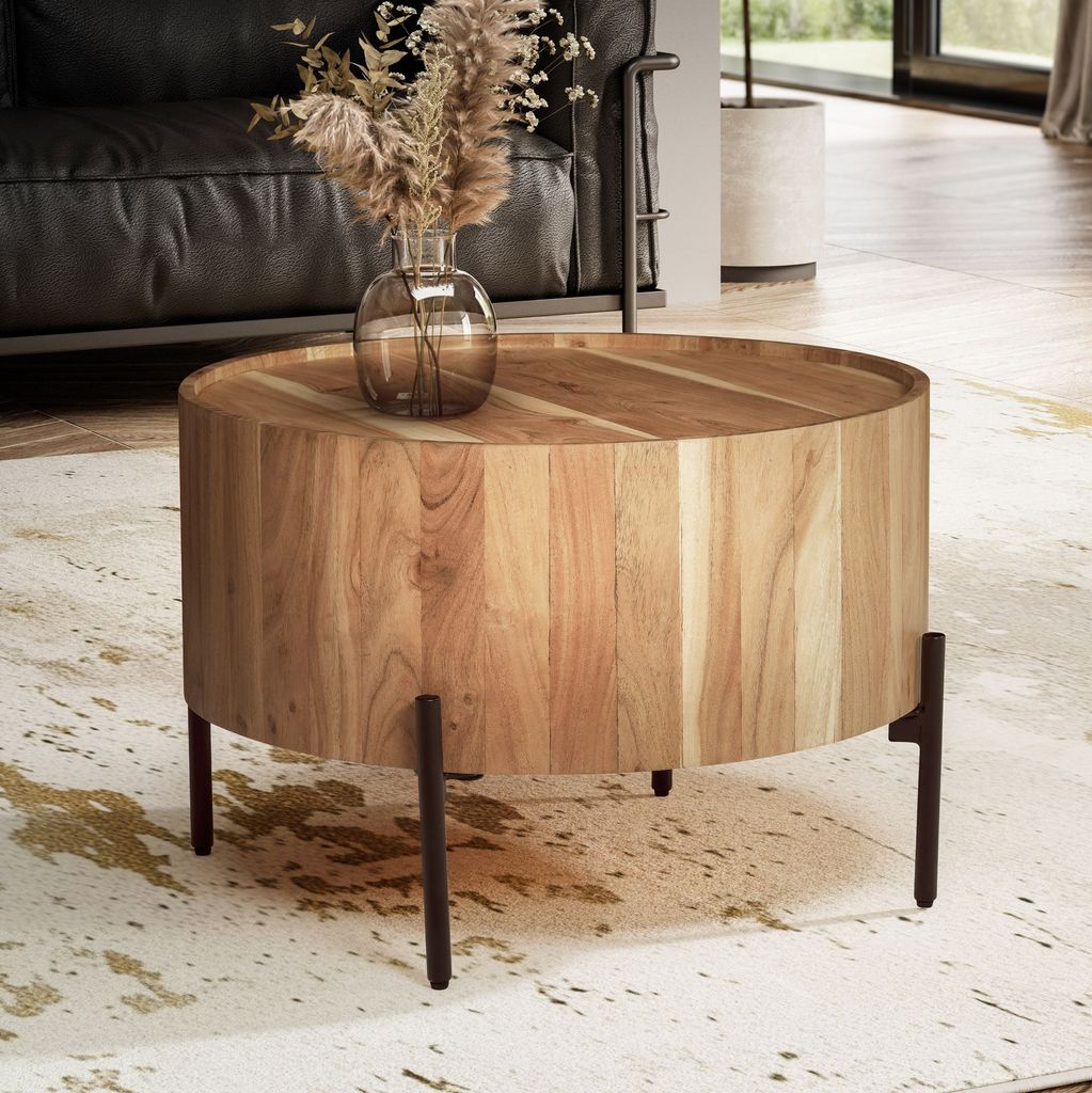 A round wooden coffee table with a vase of dried flowers on top, placed on a rug in a living room.